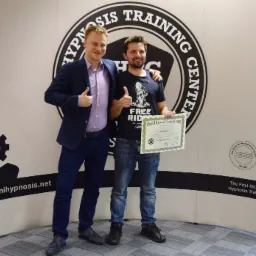 Dwaj mężczyźni uśmiechnięci, z podniesionymi kciukami, pozują na tle ścianki reklamowej Omni Hypnosis Training Center, jeden z nich trzyma certyfikat z imieniem i nazwiskiem.