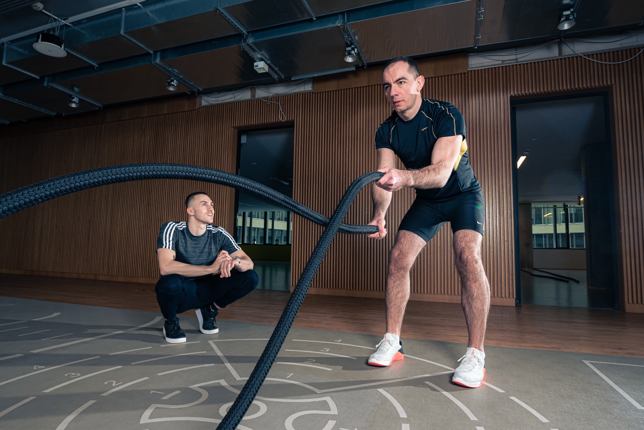 Mężczyzna w czarnym stroju sportowym ćwiczy z grubymi linami w siłowni, pod nadzorem drugiego mężczyzny w stroju Adidas.