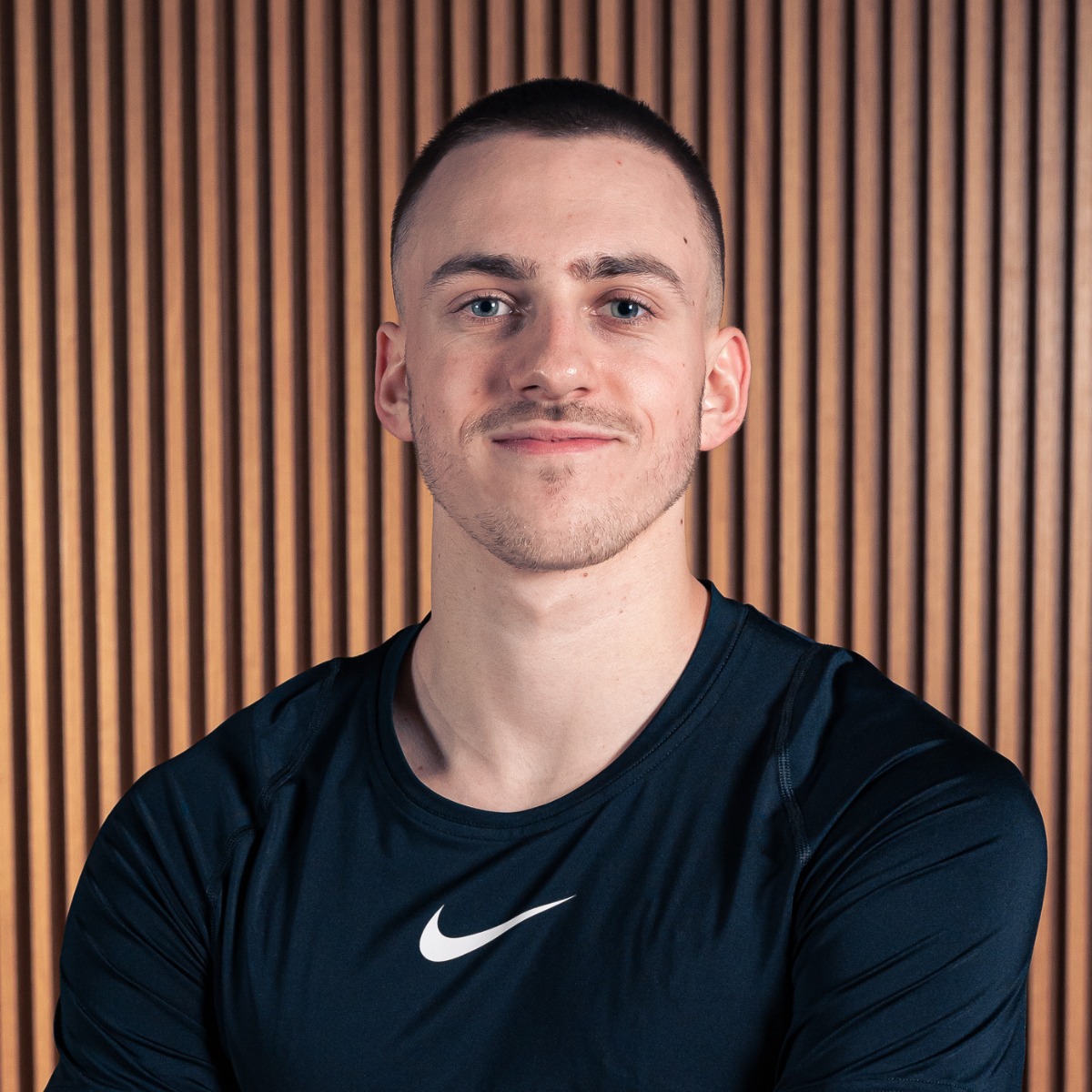 Portret młodego mężczyzny w sportowej koszulce z logo Nike na tle ściany z drewnianych paneli.