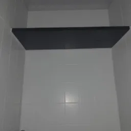 Wnęka w łazience wyłożona białymi, kwadratowymi płytkami ceramicznymi, z zamontowaną, ciemną, prostokątną półką na górze.