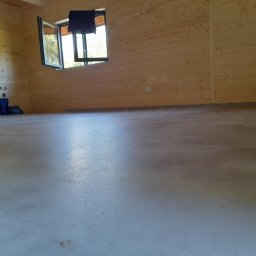 Jarosław Michoń - Surowe wnętrze z drewnianymi ścianami i szarą wylewką podłogową, otwarte okno z czarną ramą w górnej części ściany, widoczne instalacje na ścianie. Na podłodze leżą torby.