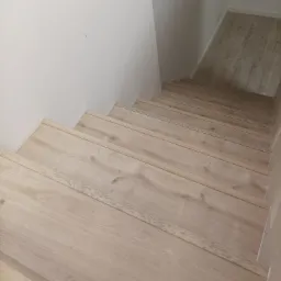 Jasne, drewniane schody z widoczną strukturą drewna, prowadzące w dół, przy białej ścianie.