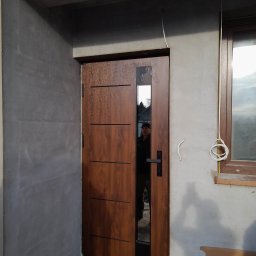 Jarosław Michoń - Brązowe drzwi wejściowe z przeszkleniem, zamontowane w surowym stanie budynku z widocznym oknem i przewodami elektrycznymi.