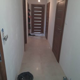 Jarosław Michoń - Wnętrze korytarza z zamontowanymi drzwiami wewnętrznymi, widoczny pracownik z narzędziami w tle, na podłodze wiadro z pozostałościami po pracach.