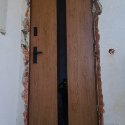 Jarosław Michoń - Drzwi wejściowe z drewnopodobną okleiną i czarnym pasem pośrodku, świeżo osadzone w surowym otworze murarskim z widocznymi cegłami i pianką montażową.