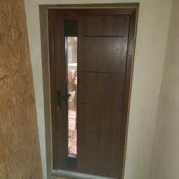 Jarosław Michoń - Nowo zamontowane, brązowe drzwi wejściowe z szybą, widoczne z bliska, z kluczem w zamku, na tle surowych ścian i płyty OSB, w trakcie prac wykończeniowych.