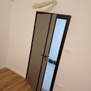 Szare drzwi wewnętrzne z czarną ramą, zamontowane w białym pomieszczeniu. Nad drzwiami widoczna owinięta folią instalacja. Podłoga z paneli drewnopodobnych.