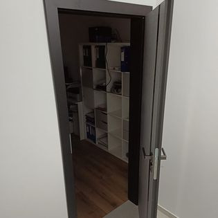 Otwarta, szara, metalowa ościeżnica z widokiem na pomieszczenie biurowe z białymi półkami i drewnianą podłogą. Widoczny fragment ściany pomalowanej na biało.