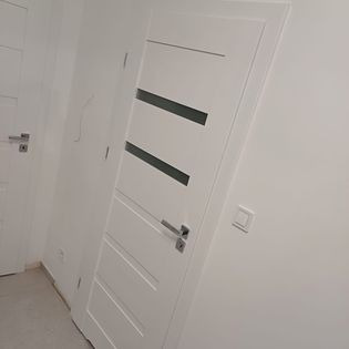 Białe drzwi wewnętrzne z trzema poziomymi, szarymi wstawkami, zamontowane w białej ścianie. Widoczna klamka i ościeżnica. Ujęcie z niskiego kąta.