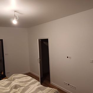 Wnętrze pokoju po świeżym pomalowaniu ścian na biało, widoczne gniazdka elektryczne, drzwi wewnętrzne i fragment łóżka z pościelą w paski.