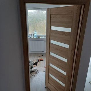 Otwarta, dębowa, wewnętrzna ościeżnica z czterema poziomymi, mlecznymi szybami, widok na pomieszczenie z grzejnikiem, oknem i narzędziami na podłodze.