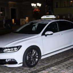 Biały samochód osobowy z lampą TAXI na dachu, zaparkowany na brukowanej ulicy w nocy, z budynkiem w tle.