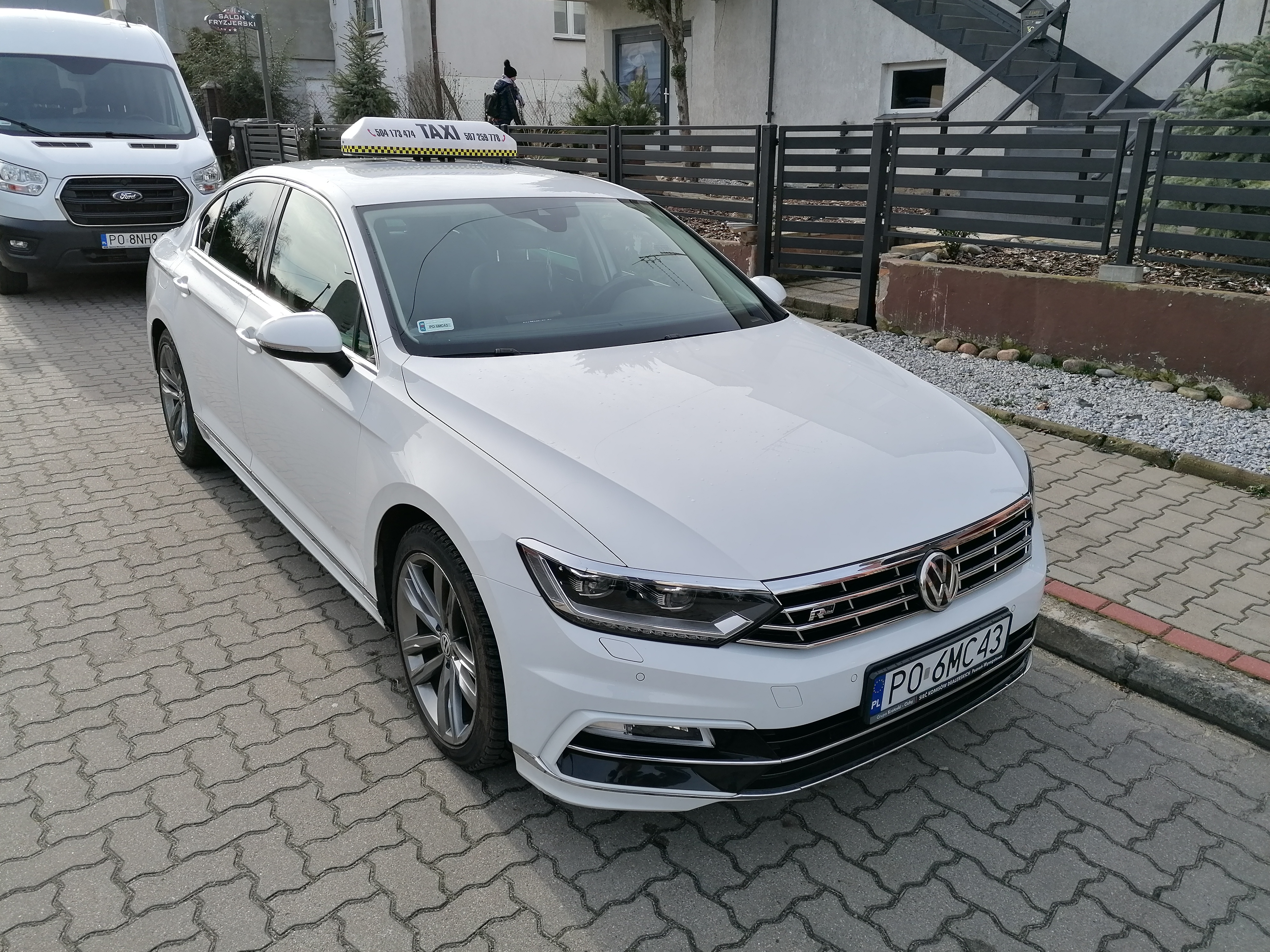 Biały samochód osobowy z napisem TAXI na dachu, zaparkowany na brukowanej ulicy przed budynkiem.