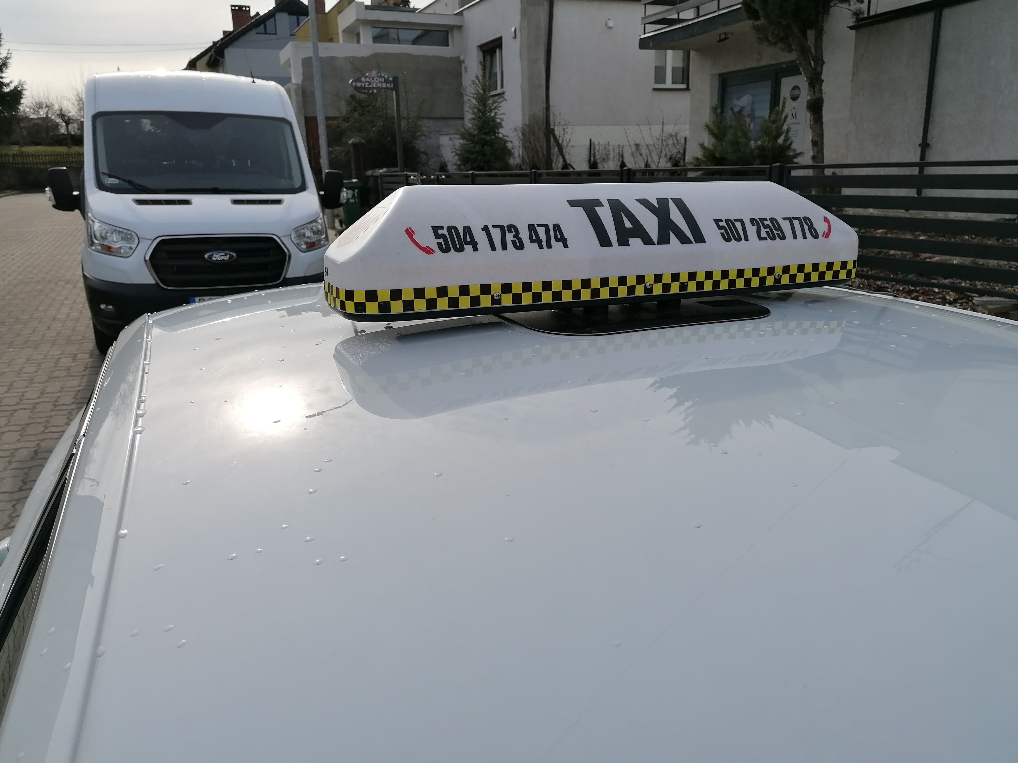 Biały samochód z lampą Taxi na dachu, widoczne numery telefonów i czarno-żółta szachownica, w tle biały bus Ford Transit.