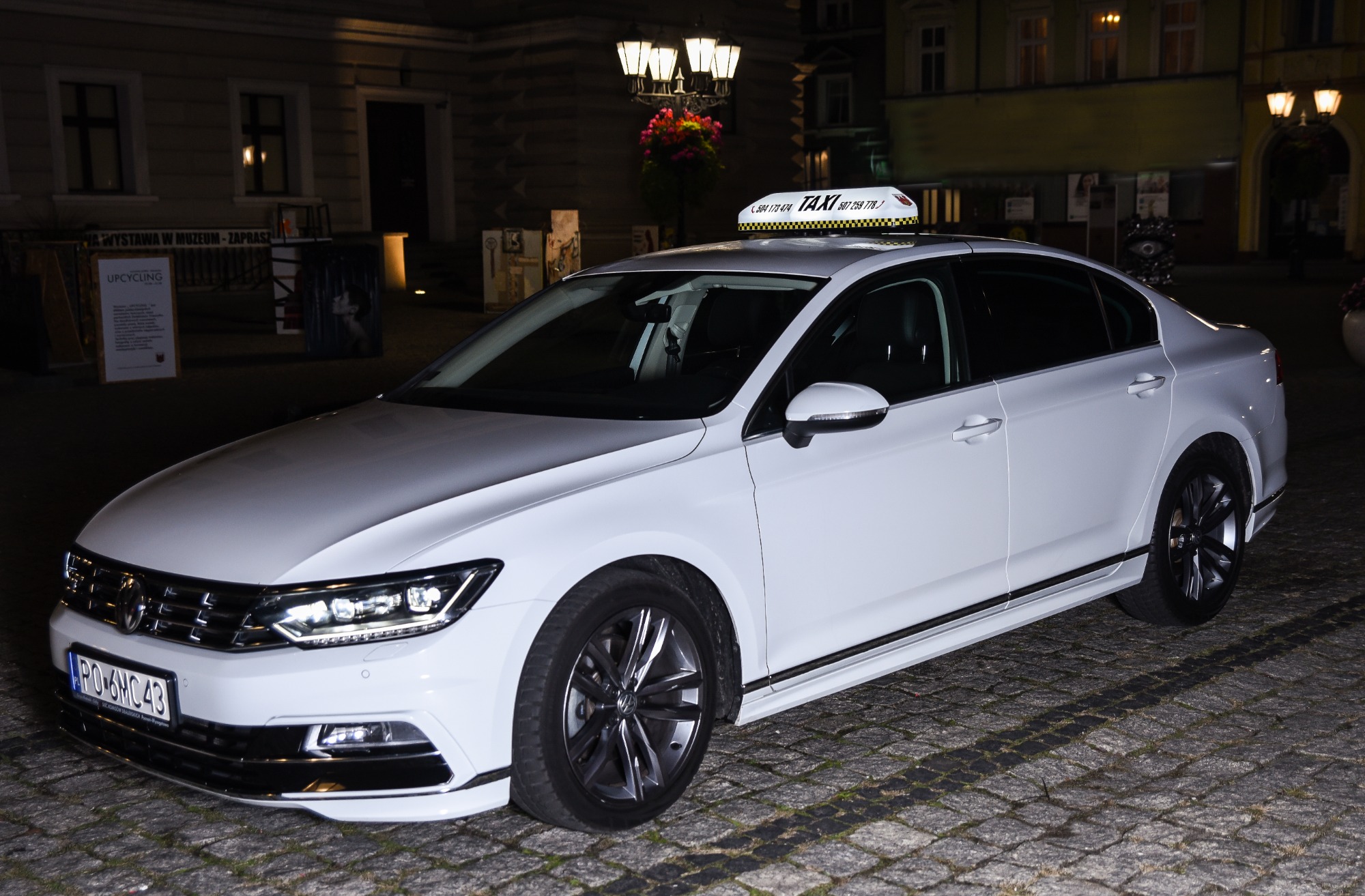 Biały samochód osobowy z lampą TAXI na dachu, zaparkowany na brukowanej ulicy w nocy, z budynkiem w tle.