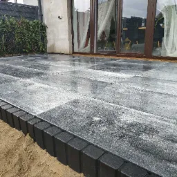 Świeżo ułożony, mokry taras z szarych, prostokątnych płyt betonowych, obrzeżony ciemną kostką, z widoczną warstwą piasku i przesuwne drzwi tarasowe w tle.