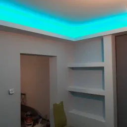 Wykończone wnętrze z podwieszonym sufitem z oświetleniem LED w kolorze turkusowym i wnękami ściennymi, widoczne farby i narzędzia malarskie oraz krzesło w tle.