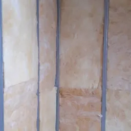 Montaż izolacji z wełny mineralnej w metalowym stelażu ścianki działowej, widoczne łączenia płyt i niedoskonałości w wypełnieniu przestrzeni.