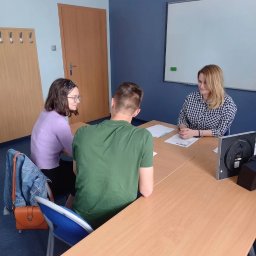 Nova Centrum - Szkoła Języków Obcych - Nauczycielka prowadzi lekcję w klasie dla dwóch uczniów. Widoczna tablica, projektor i biurko. Uczniowie słuchają nauczycielki. Sala jest jasna i przestronna.