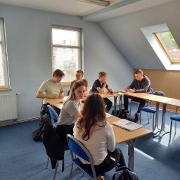 Nova Centrum - Szkoła Języków Obcych - Lekcja w jasnej sali: uczniowie siedzą przy biurkach, rozmawiają, pracują z materiałami. Widoczne okna, niebieska wykładzina i białe ściany. Lekcja w toku.