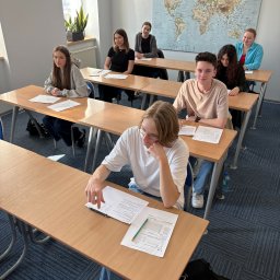 Nova Centrum - Szkoła Języków Obcych - Sala lekcyjna z uczniami siedzącymi przy biurkach, mapa świata na ścianie, naturalne światło z okna. Jeden uczeń rozwiązuje zadanie.