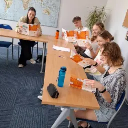 Lekcja języka: uczniowie czytają podręczniki przy stole w klasie. Na ścianie mapa świata, butelki z wodą i piórnik na blacie. Jasne, naturalne światło.