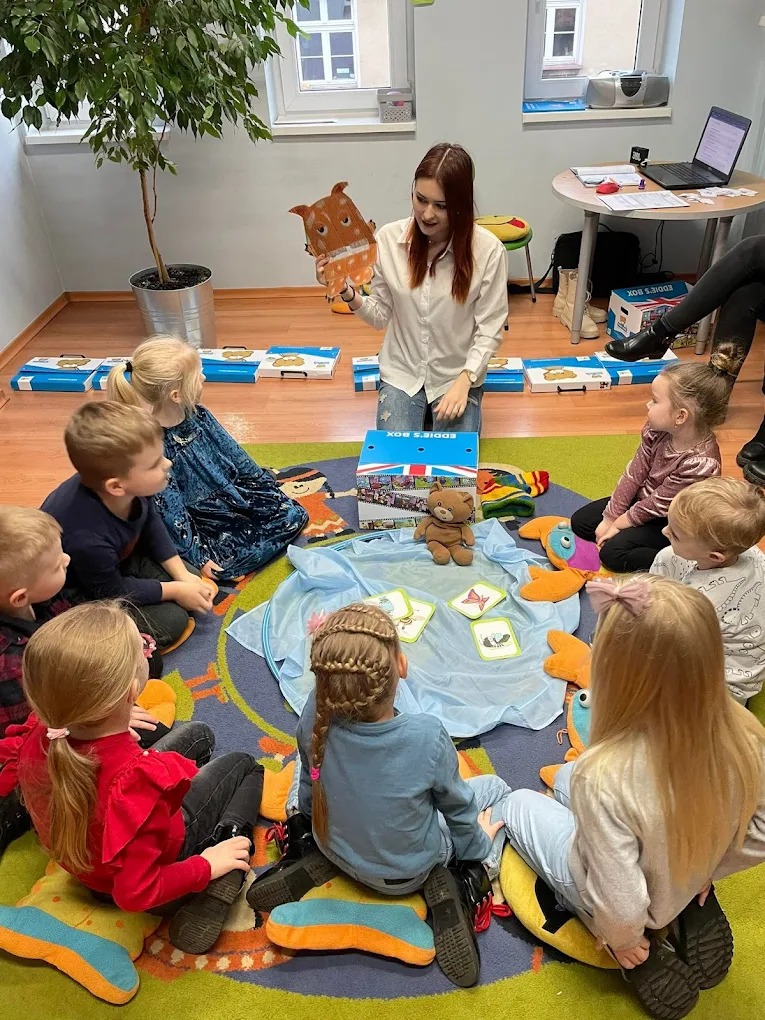 Nauczycielka prezentuje kartonową sowę dzieciom siedzącym w kręgu na kolorowym dywanie, podczas zajęć w szkole językowej. W tle widoczne pudełka i laptop.