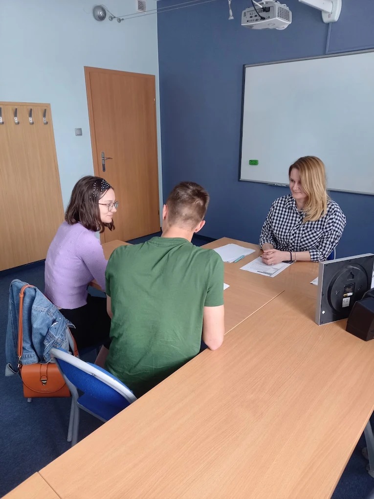 Nauczycielka prowadzi lekcję w klasie dla dwóch uczniów. Widoczna tablica, projektor i biurko. Uczniowie słuchają nauczycielki. Sala jest jasna i przestronna.