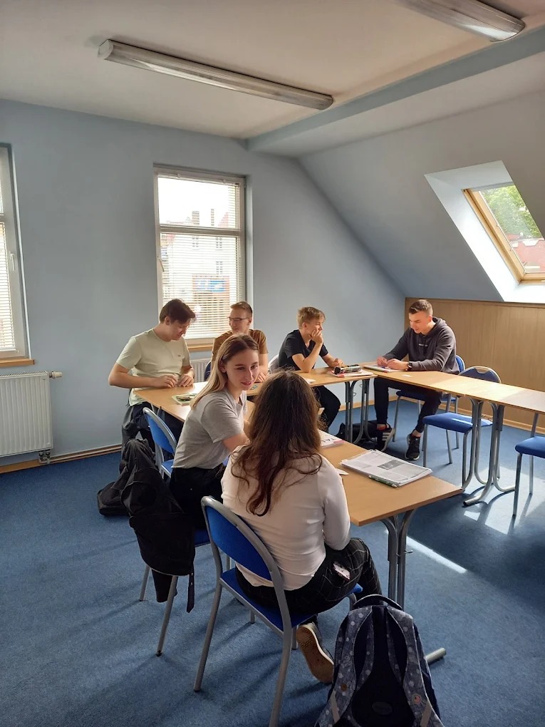 Lekcja w jasnej sali: uczniowie siedzą przy biurkach, rozmawiają, pracują z materiałami. Widoczne okna, niebieska wykładzina i białe ściany. Lekcja w toku.