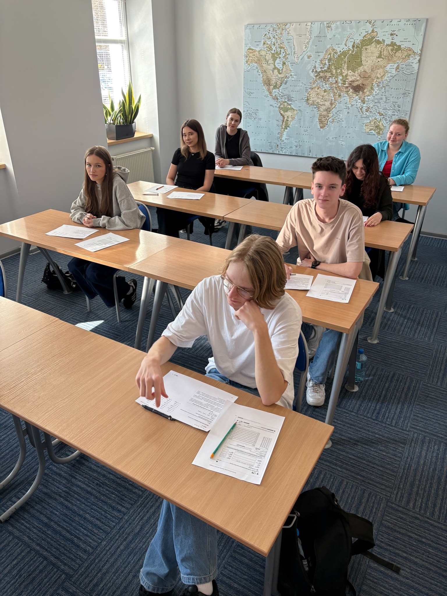 Sala lekcyjna z uczniami siedzącymi przy biurkach, mapa świata na ścianie, naturalne światło z okna. Jeden uczeń rozwiązuje zadanie.