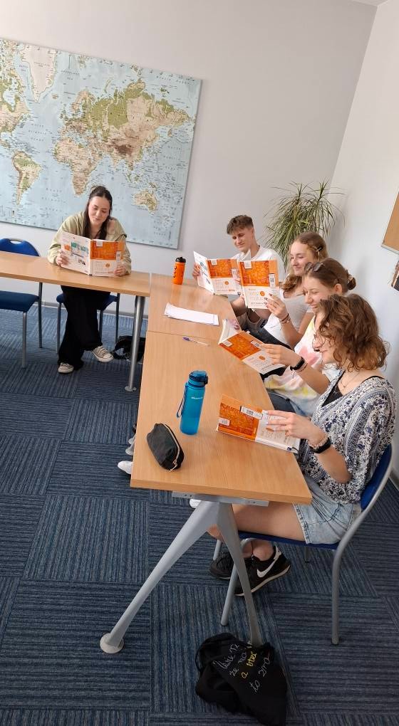 Lekcja języka: uczniowie czytają podręczniki przy stole w klasie. Na ścianie mapa świata, butelki z wodą i piórnik na blacie. Jasne, naturalne światło.