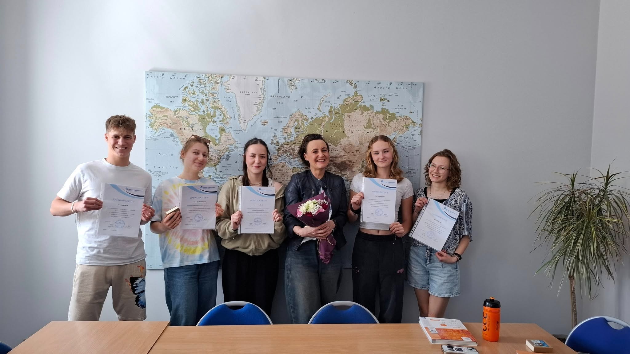 Wręczenie certyfikatów ukończenia kursu językowego grupie uczniów z nauczycielem na tle mapy świata w jasnym pomieszczeniu.
