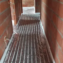 Widok na etapie montażu ogrzewania podłogowego w surowym wnętrzu z czerwonej cegły, rury grzewcze ułożone na folii izolacyjnej z siatką.