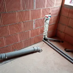 Fragment instalacji wodnej w surowym pomieszczeniu z ceglanymi ścianami, widoczne rury PCV, pianka montażowa wokół przyłączy i otwory w posadzce.