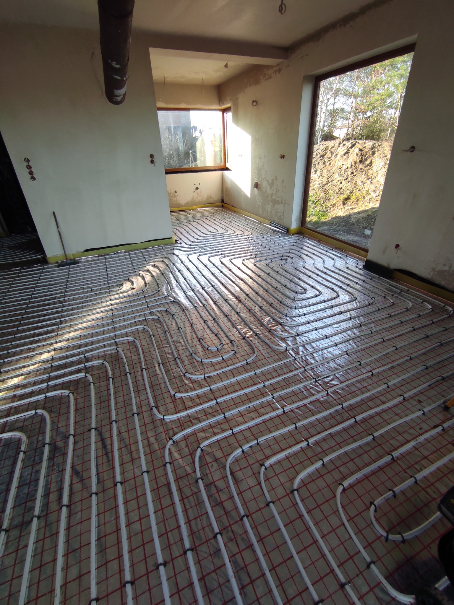 Białe rurki ogrzewania podłogowego ułożone w pętle na folii z czerwoną kratką w surowym wnętrzu, widoczne okna z widokiem na zewnątrz, instalacja w trakcie realizacji.