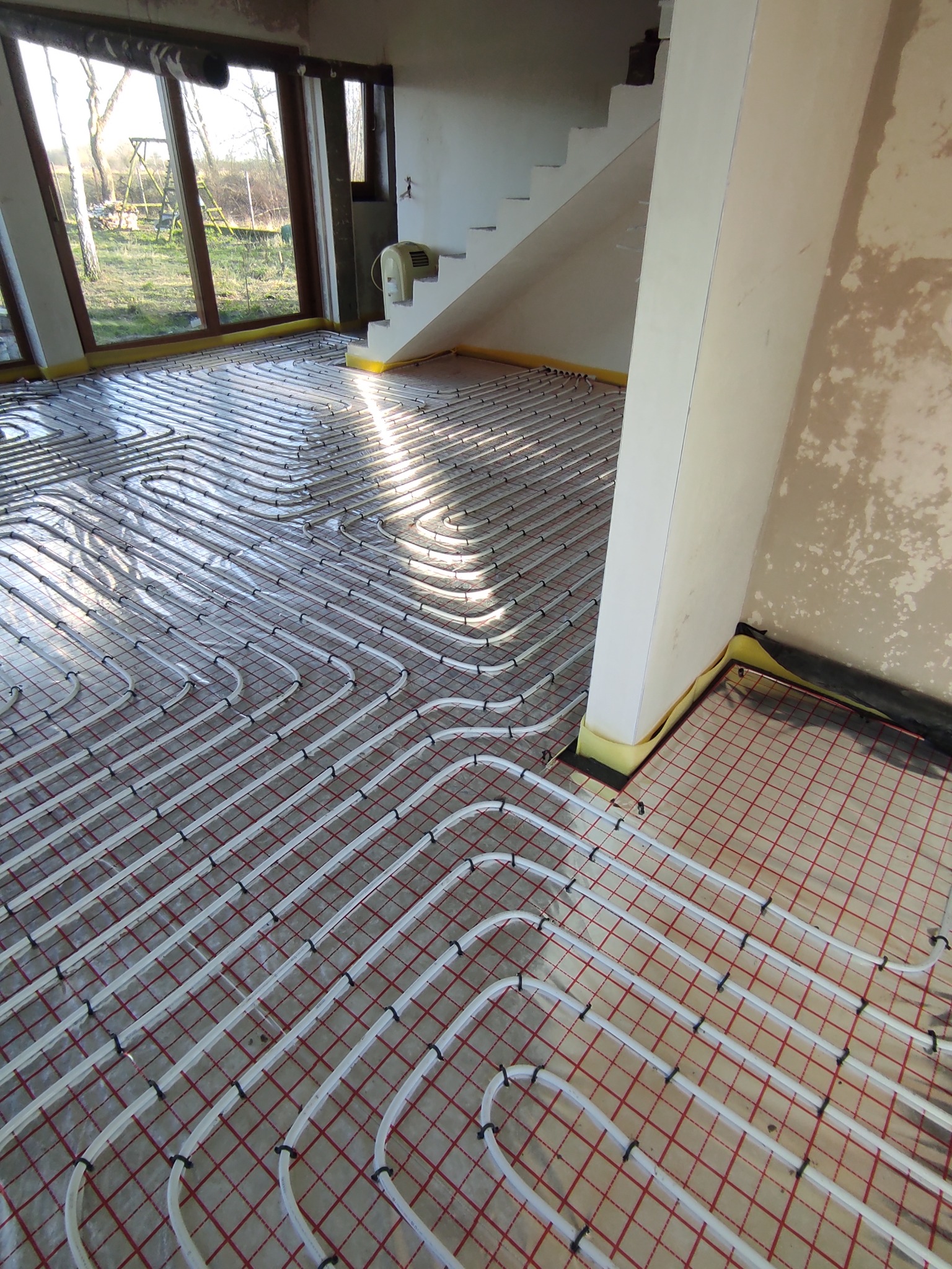 Białe rury ogrzewania podłogowego ułożone spiralnie na czerwonej siatce w surowym wnętrzu domu w trakcie budowy, widoczne okno i schody.