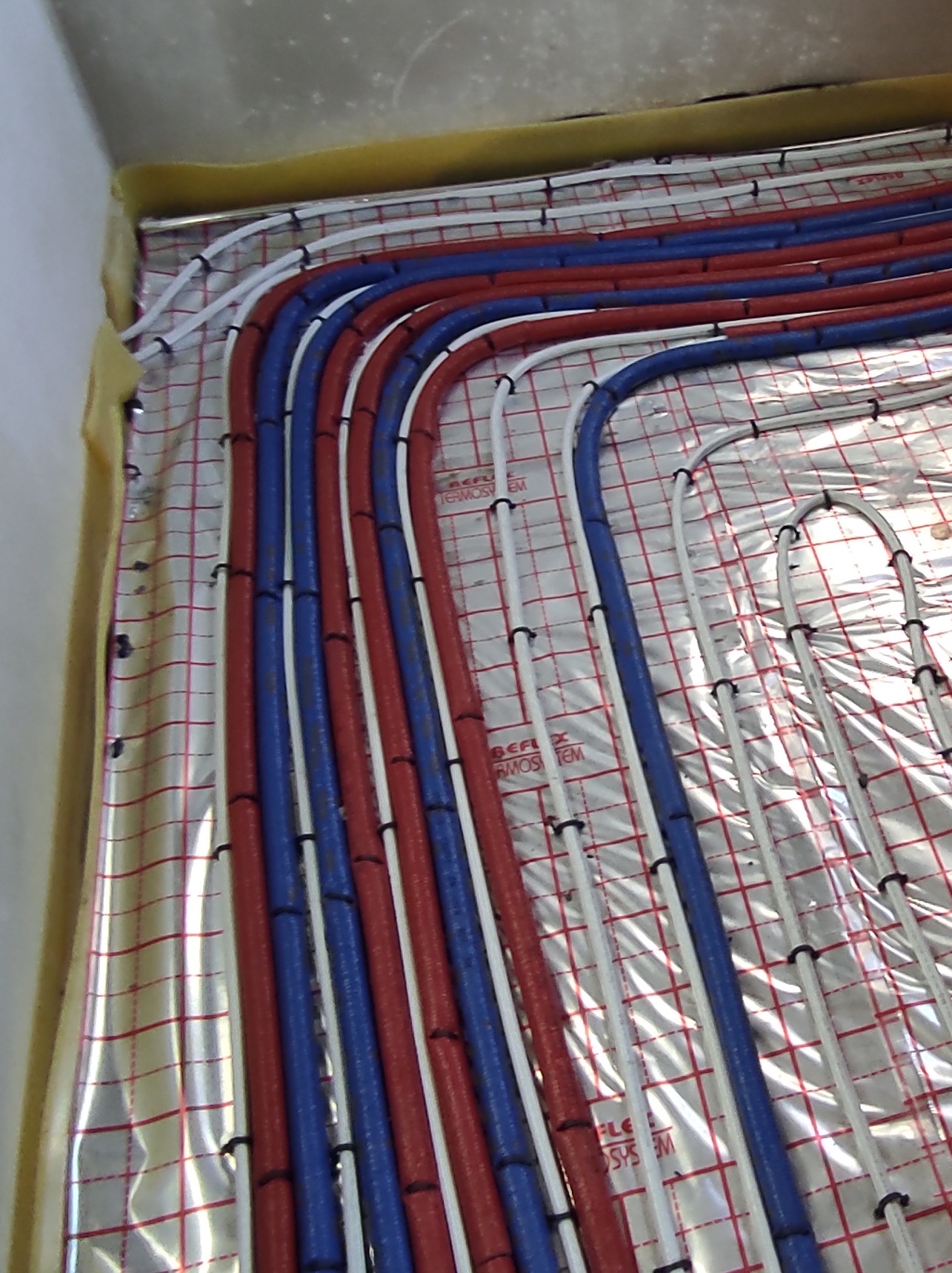 Rury grzewcze podłogowe w kolorach czerwonym, niebieskim i białym, zamocowane na folii refleksyjnej z siatką, widoczne czarne opaski mocujące.