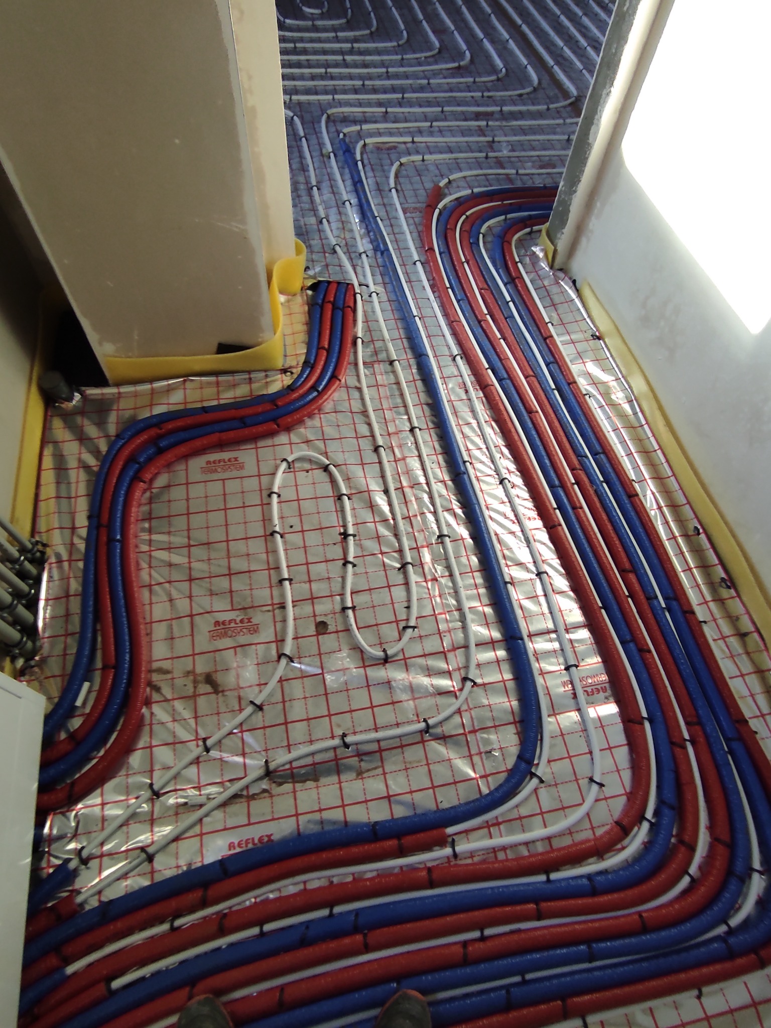 Widok z góry na rozłożone rury ogrzewania podłogowego w kolorach czerwonym i niebieskim, przymocowane do folii z siatką, widoczne fragmenty ścian i podłogi.