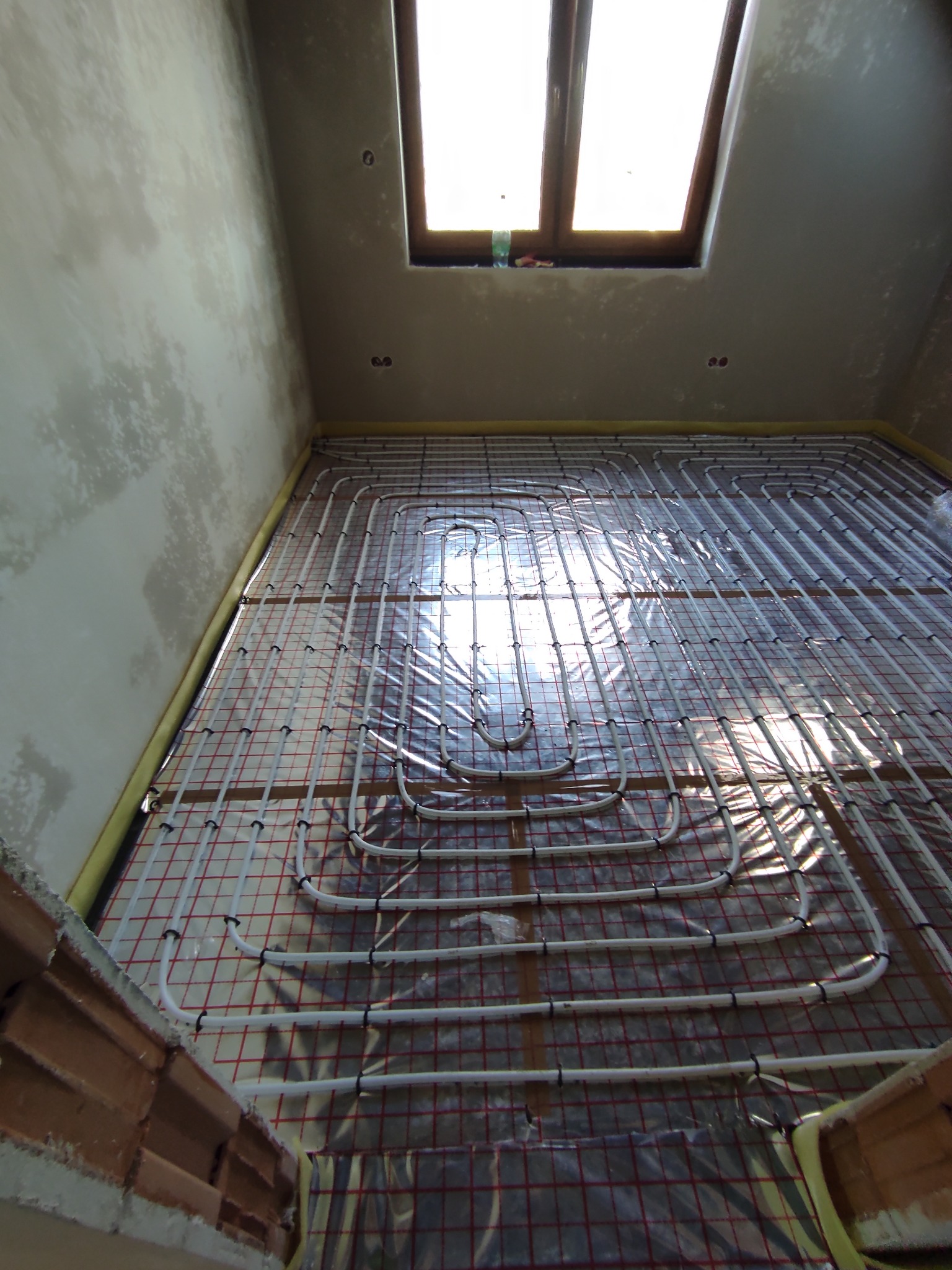 Układ rur ogrzewania podłogowego na folii aluminiowej z siatką w surowym wnętrzu, widok z góry na instalację przed wylewką, okno dachowe w tle.