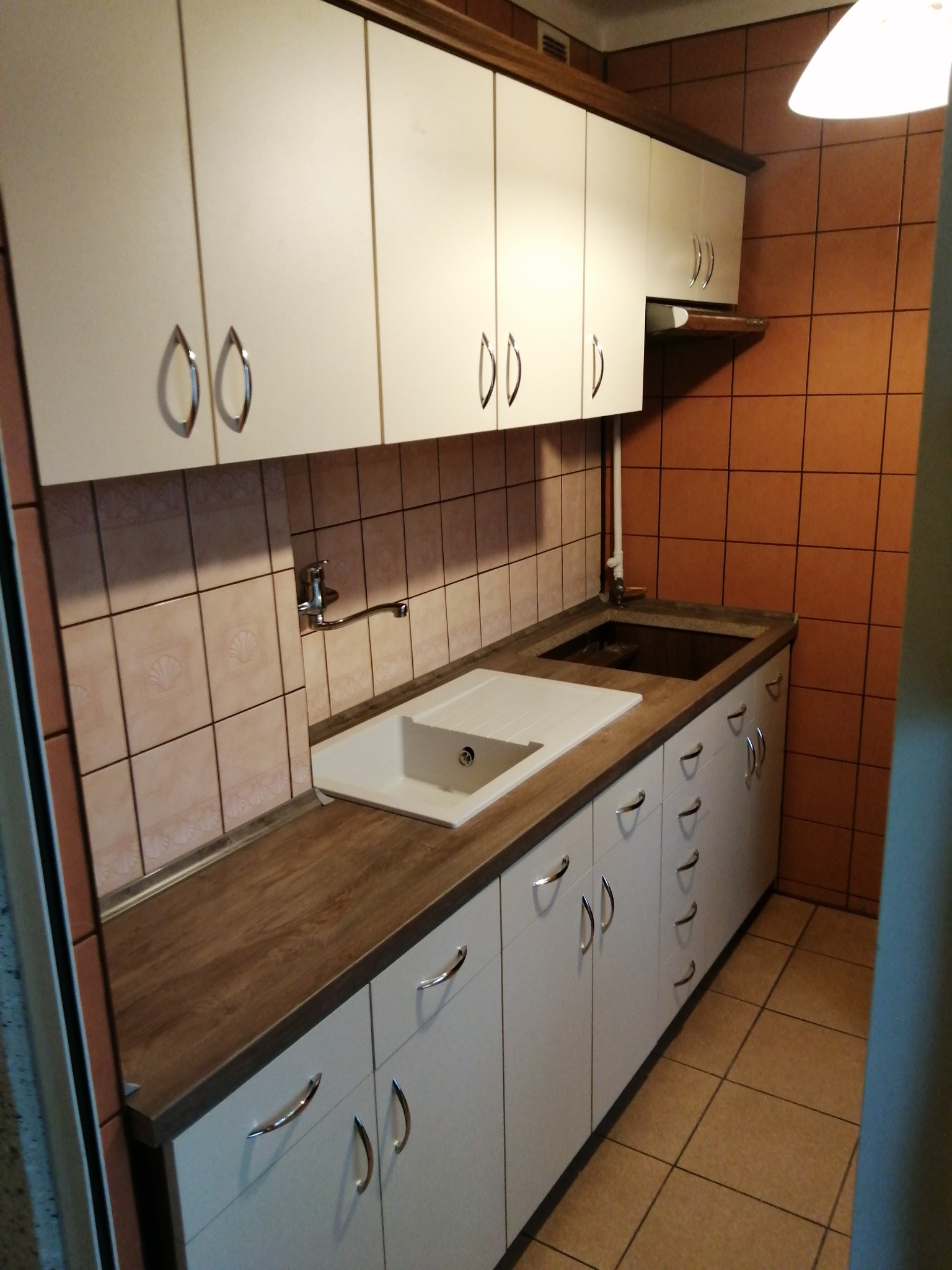 Kuchnia w Skawinkach z białymi szafkami z srebrnymi uchwytami i drewnianym blatem, widoczna biała umywalka i bateria, brązowe płytki na ścianie.