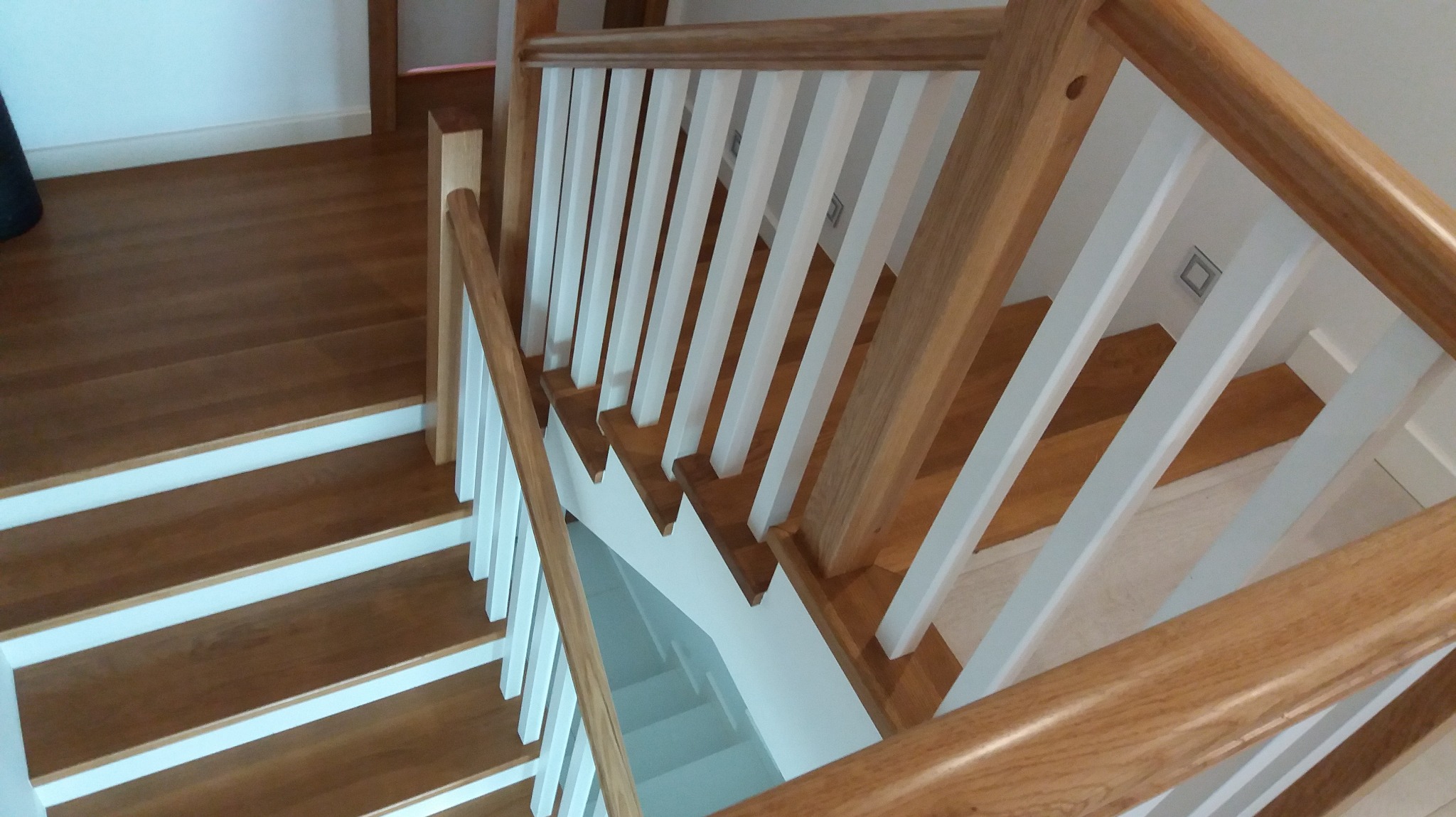 Drewniane schody z białymi podstopnicami i balustradą z jasnego drewna, widok z góry, oświetlenie punktowe na suficie. Detal wykończenia wnętrza.