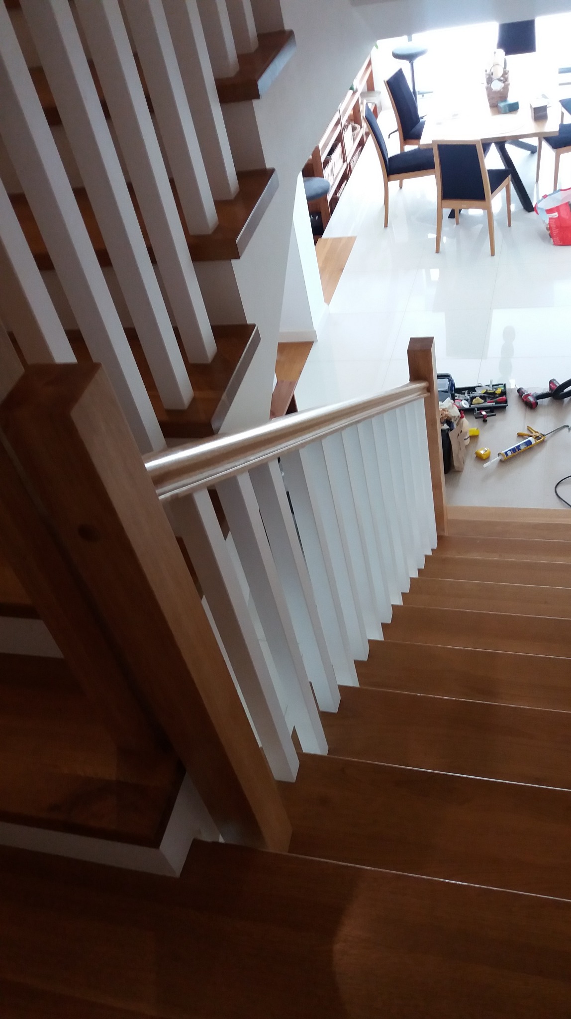 Drewniane schody z białymi balustradami, widok z góry. Na podłodze widoczne narzędzia, sugerujące trwający montaż. W tle jasne pomieszczenie z jadalnią.