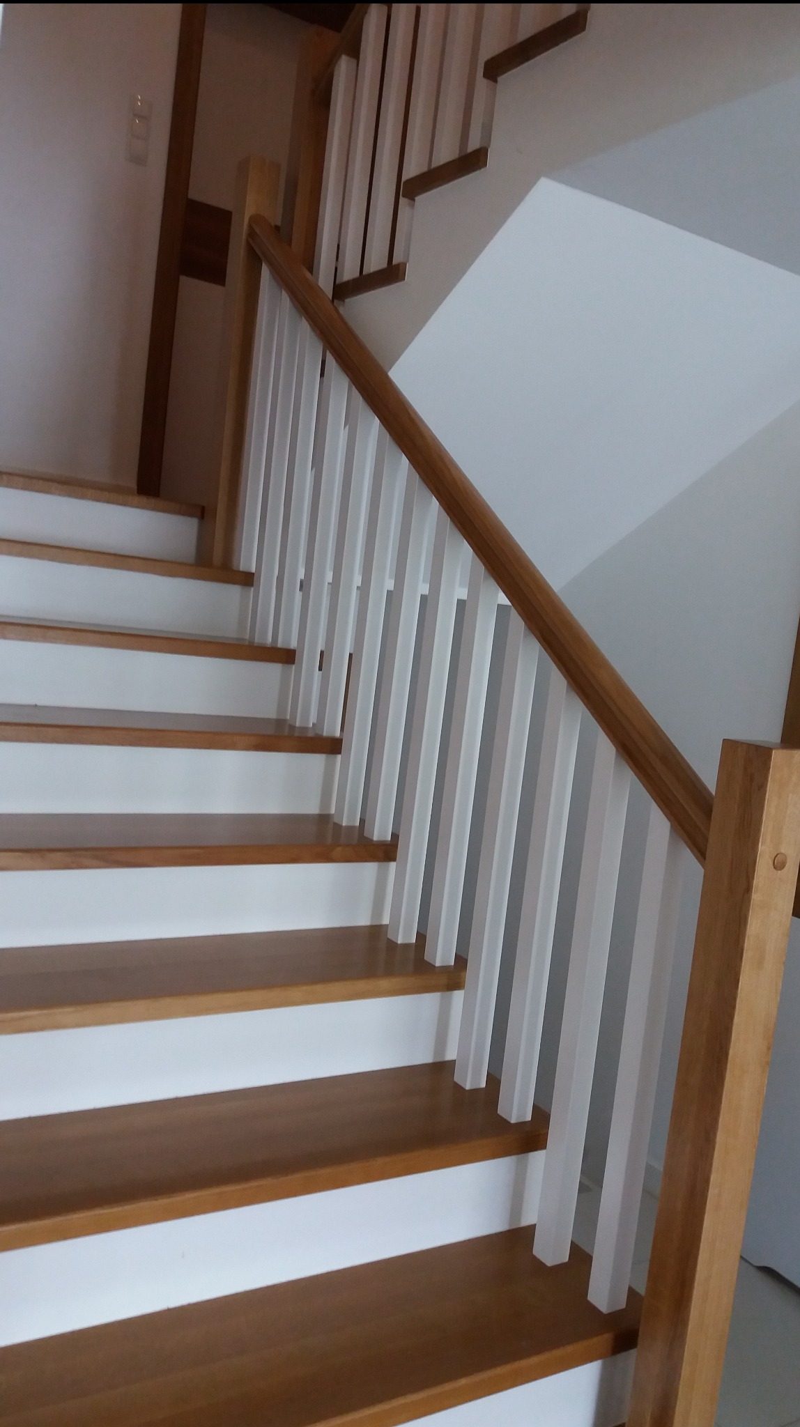 Drewniane schody z białymi balustradami, widok z dołu, ukazujący detale wykończenia i naturalne oświetlenie. Jasne drewno stopni kontrastuje z bielą.