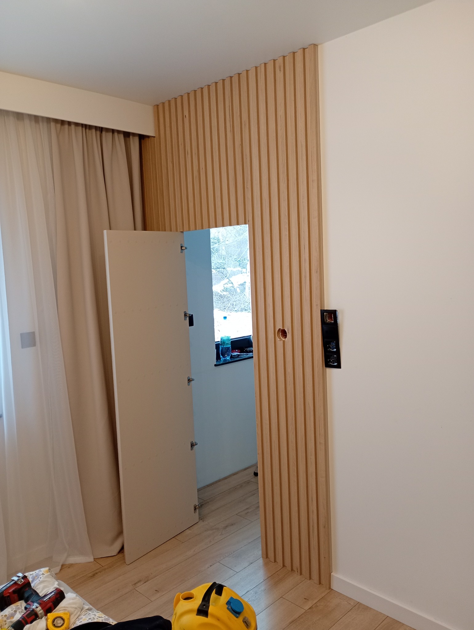 Drewniana ściana z ukrytymi drzwiami w trakcie montażu, widoczne narzędzia i niedokończone wykończenie, jasne wnętrze z oknem w tle.