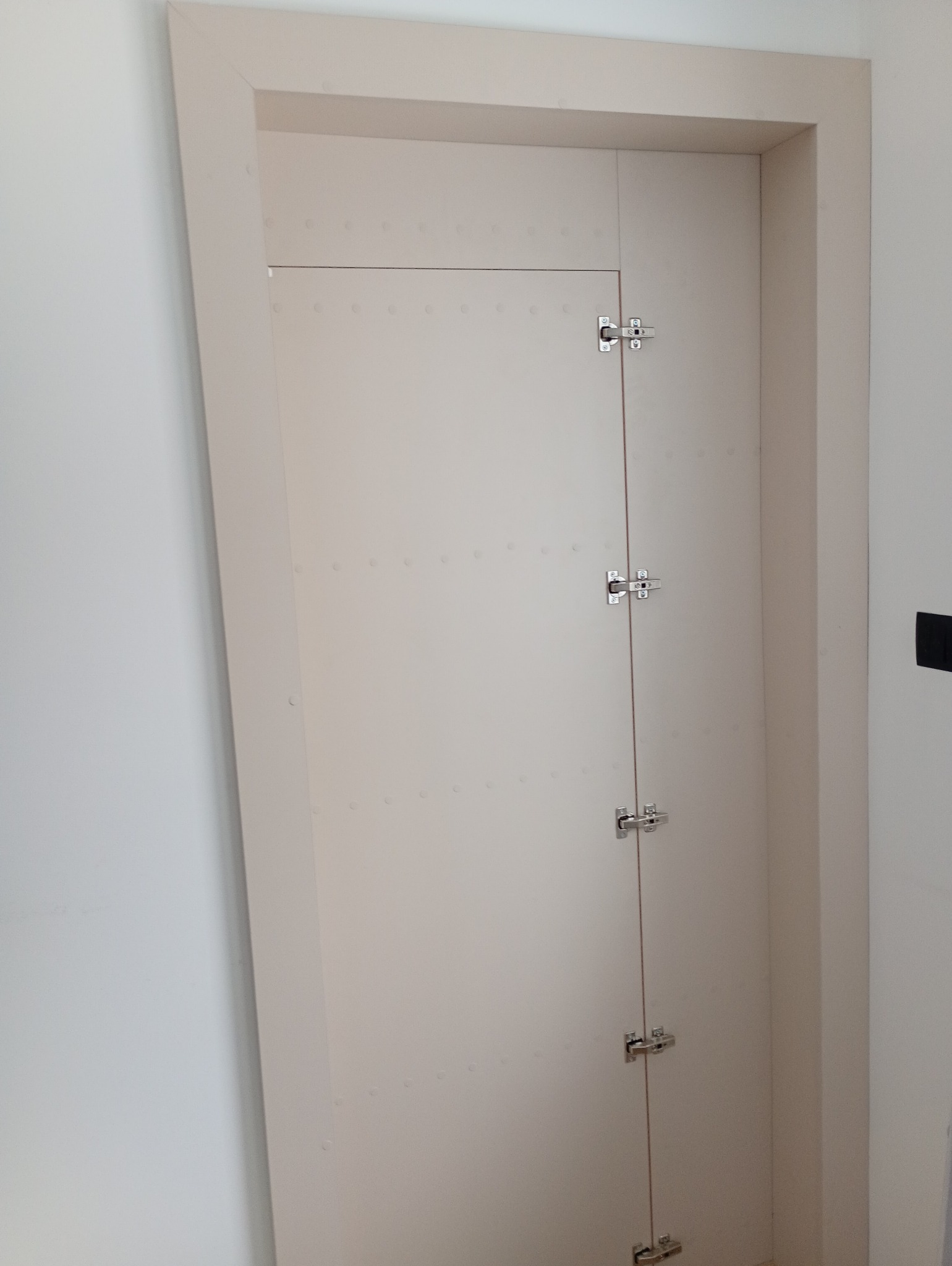 Biała, wbudowana szafa wnękowa z ukrytymi drzwiami, zawiasami i delikatnymi zdobieniami w postaci małych, okrągłych wgłębień. Minimalistyczny design.