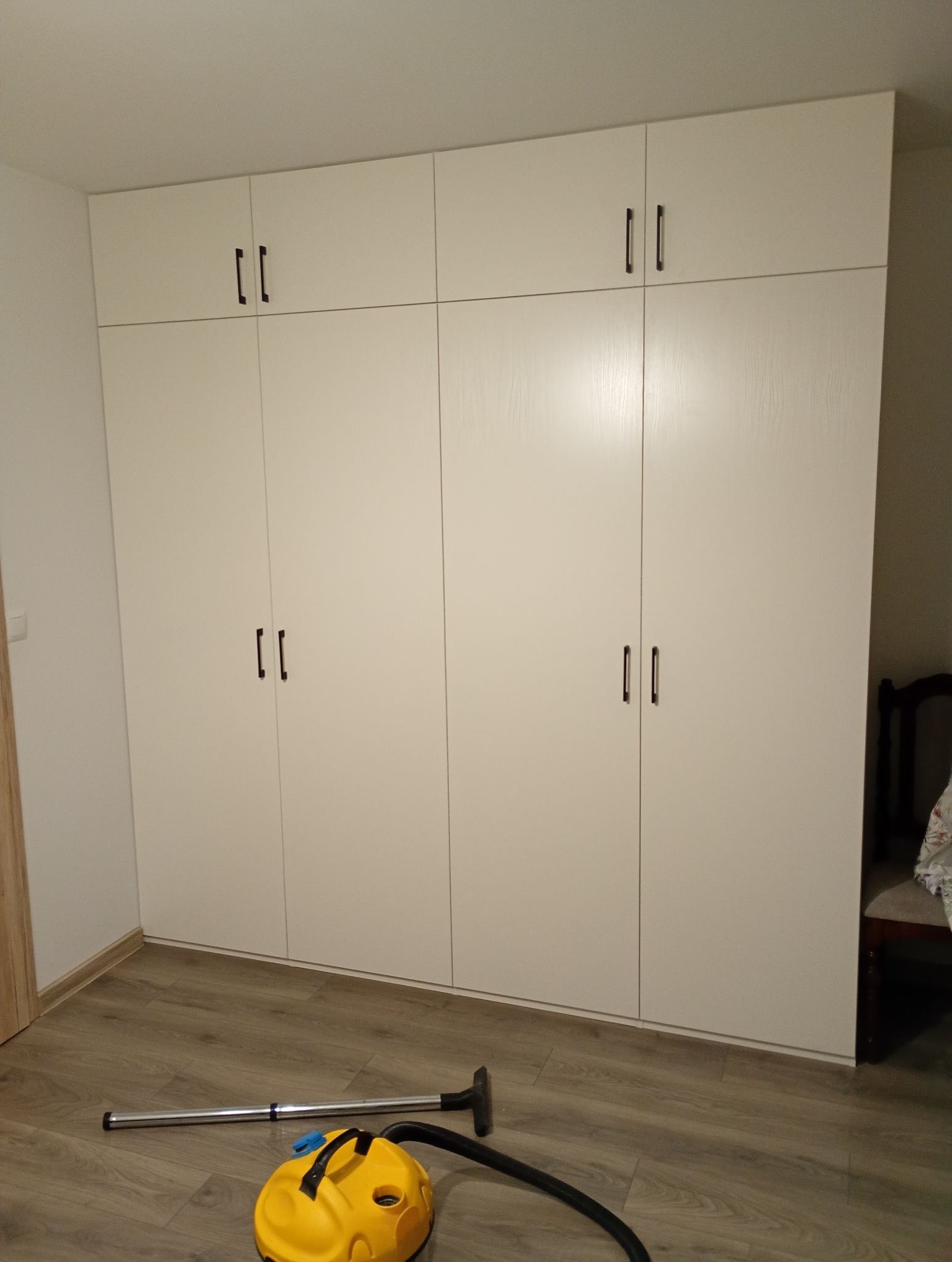 Biała szafa wnękowa z ciemnymi uchwytami, sięgająca sufitu, na podłodze odkurzacz. Minimalistyczny design, proste linie, funkcjonalne rozwiązanie do przechowywania.