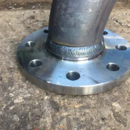 Spoina spawalnicza łącząca rurę ze stalową flanszą, widoczne otwory na śruby, szary beton w tle.