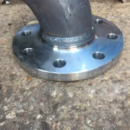 Spoina spawalnicza łącząca kolanko z kołnierzem stalowym, widoczne ślady spawania i oznaczenia na kołnierzu, powierzchnia betonowa w tle.