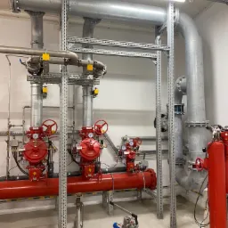 System rur stalowych, zaworów i manometrów, pomalowany na czerwono i szaro, zamontowany wewnątrz budynku, podtrzymywany przez konstrukcję wsporczą z perforowanych profili stalowych.