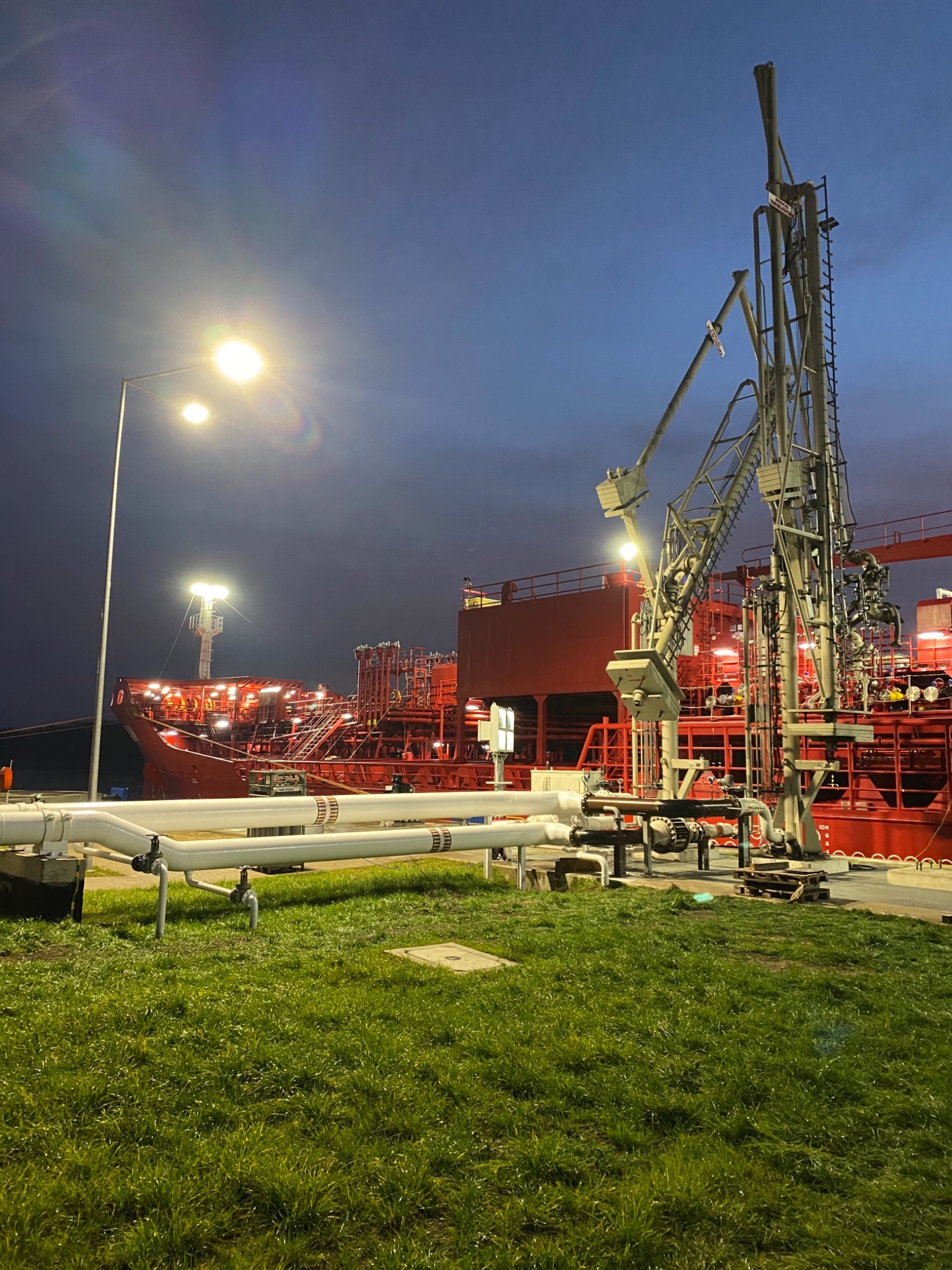 Czerwony tankowiec przy nabrzeżu w nocy, widoczny system rurociągów i pomost załadunkowy, oświetlony przez latarnie.